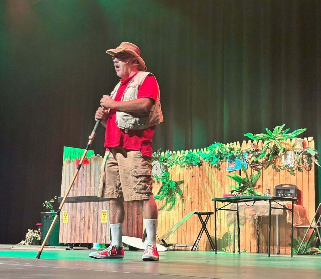 A man dressed in safari gear speaks. In the background is a fence with dense vegetation growing around it.