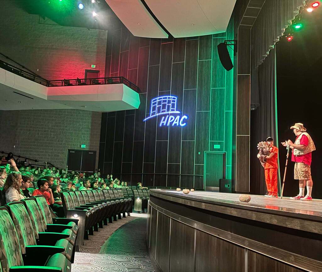 Two individuals stand on a stage while kids sit in auditorium chairs and look on.