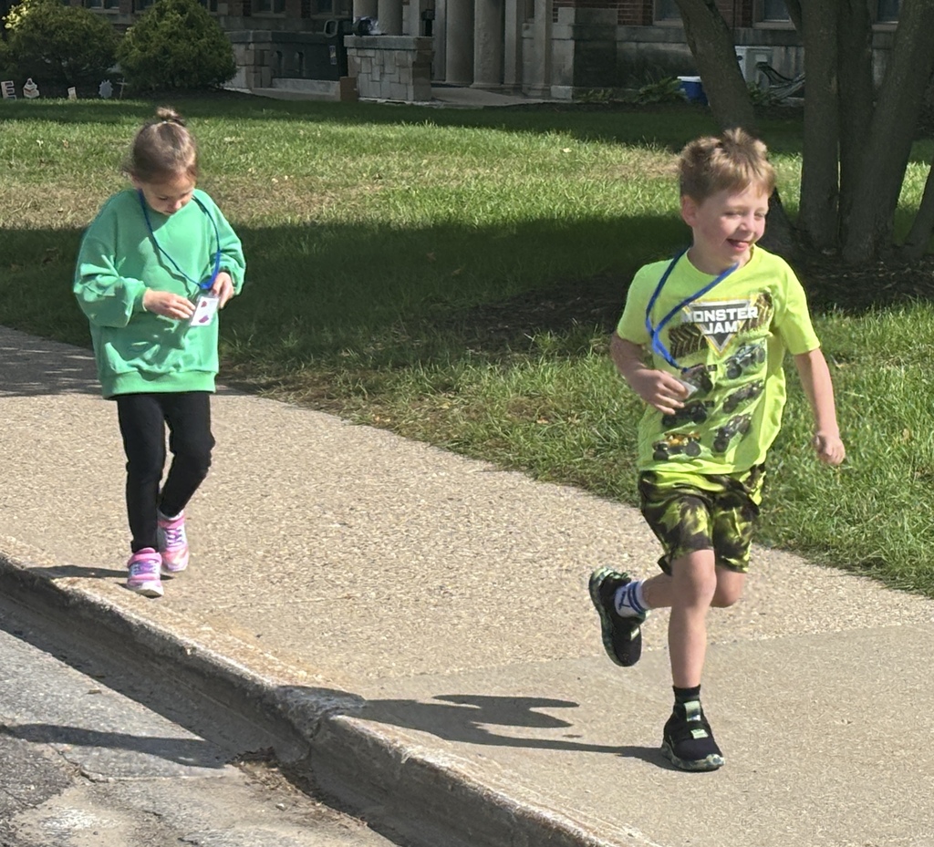 A boy in a green shirt runs down a sidewalk with a girl in a green sweatshirt walking behind.