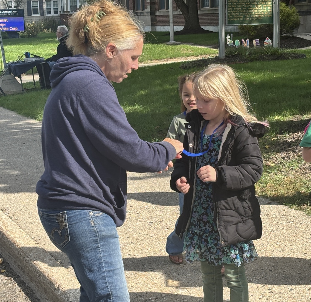A woman stands in front of a young girl, examining something that is hanging around her neck like a necklace.