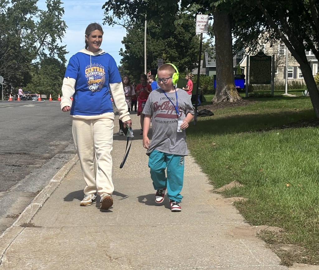 A woman in a blue shirt with a white sweatshirt layered underneath laughs with a boy in a gray shirt as they walk side-by-side down the sidewalk.