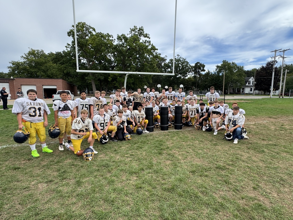 An entire youth football team with white jerseys and gold pants pose for the camera with three padded tackling dummies propped up in the center.