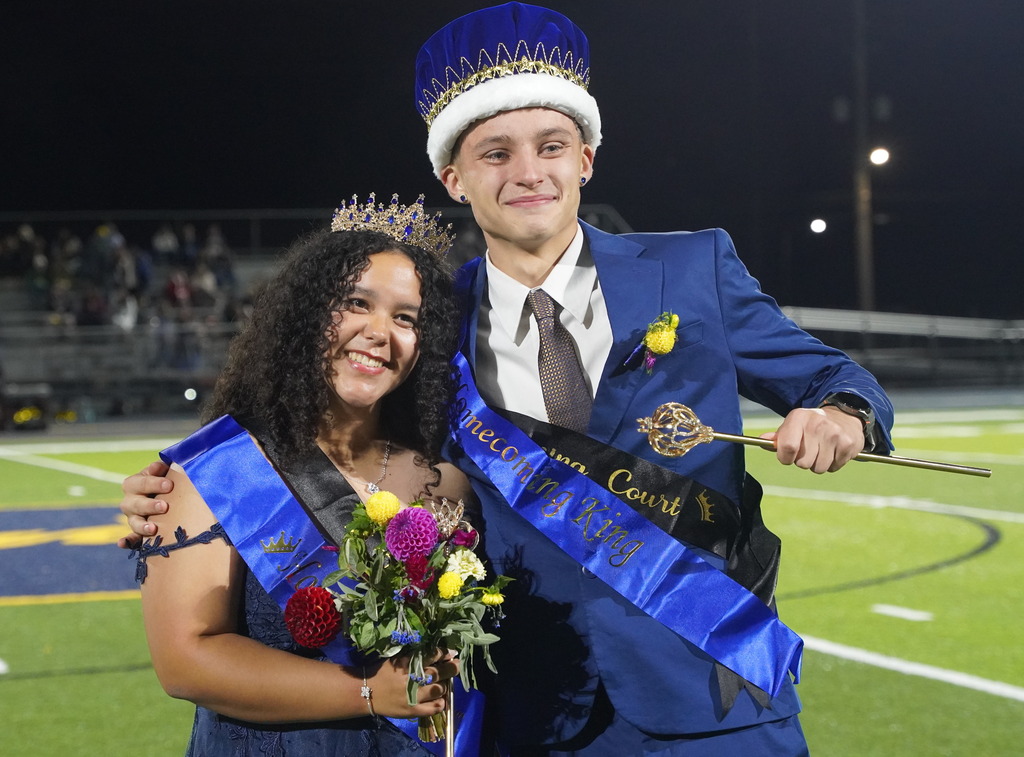 Two high school students stand on a football field with sashes across their chests and crowns on their heads while smiling