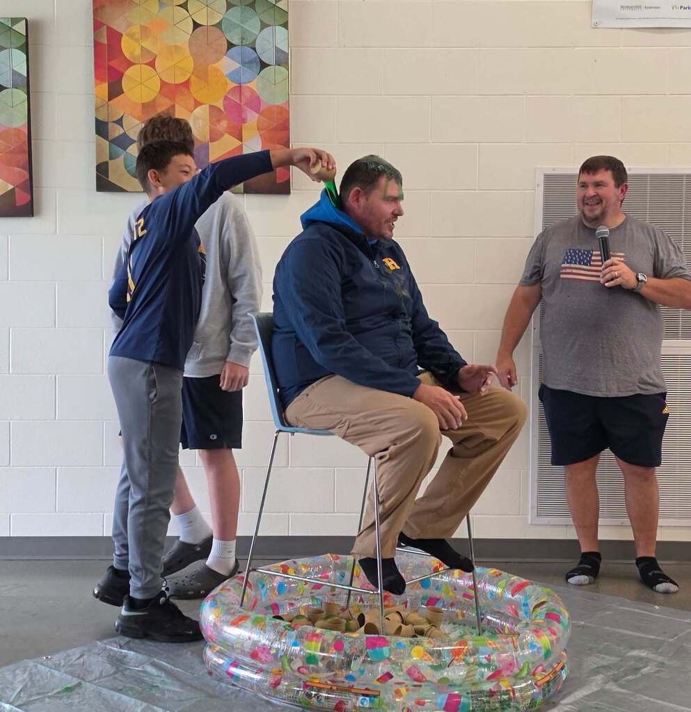 A mean weawring a blue jacket with a hood sits in a chair as a boy dumps slime from a cup on his head.