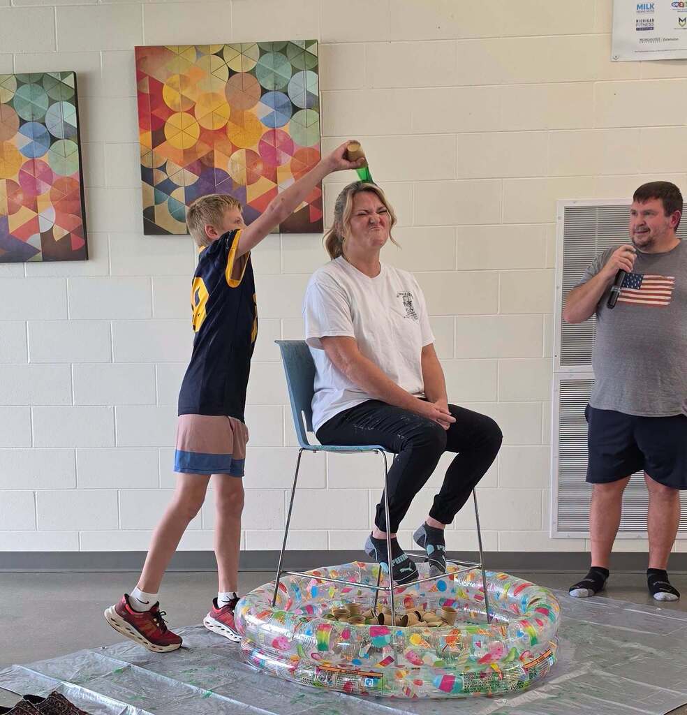 An adult female sits in a chair as a kid dumps a green substance from a cup on her head.