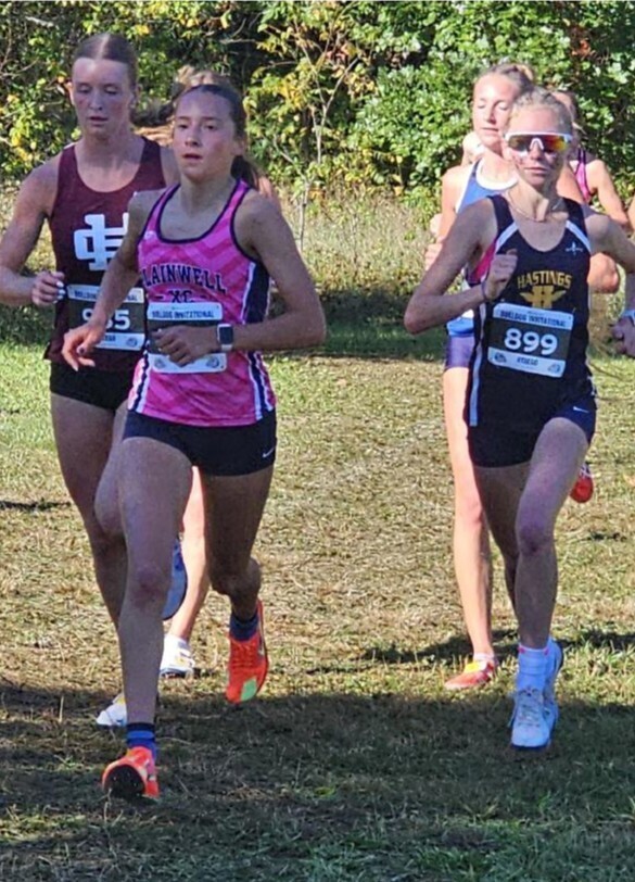 A pack of girls wearing jersey tanktops run with grass and trees in the background