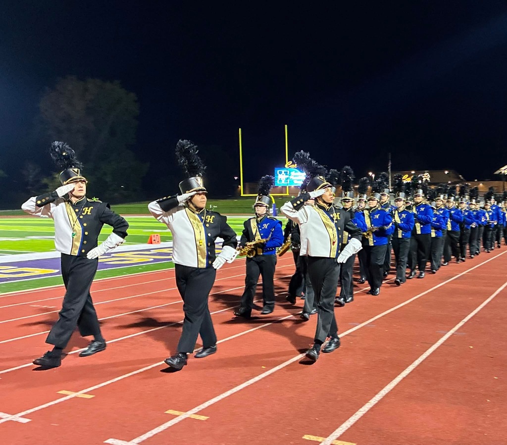 Three marching band drum majors walk down a track and field track and turn to salute. Behind is a line of blue-clad marching band members holding their various instruments.