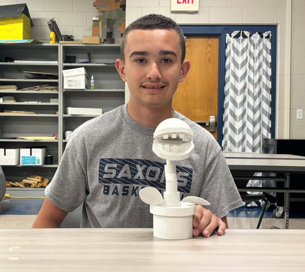 A boy in a gray t-shirt sits at a table smiling. In front of him is a white figure, resembling a character from the Super Mario franchise.