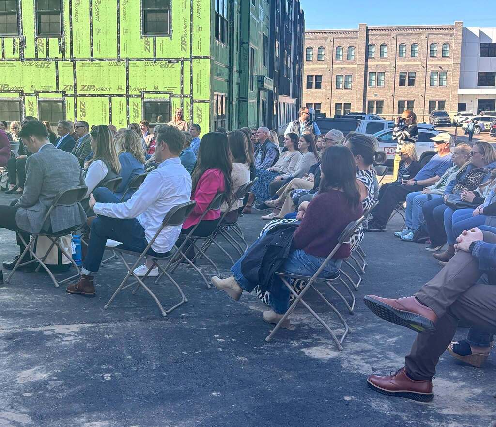 Dozens of people are seated in folding chairs outside looking ahead at a speaker who is out of frame.