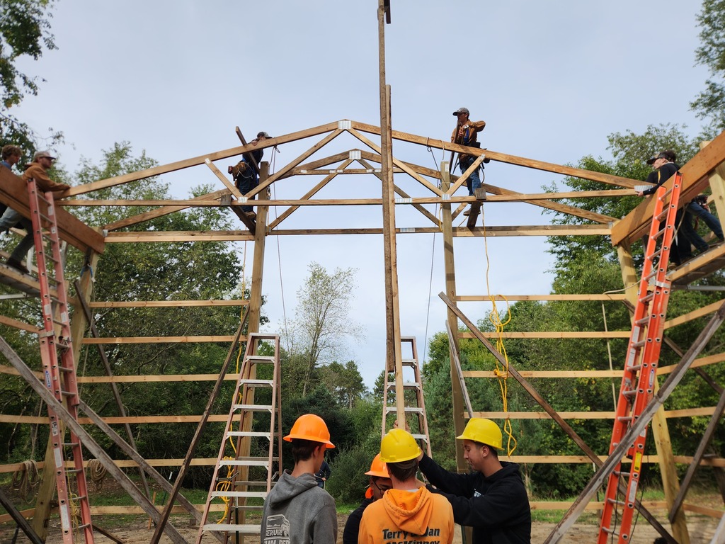 A group of young men wearing hard hats work on and around the frame of a large barn.