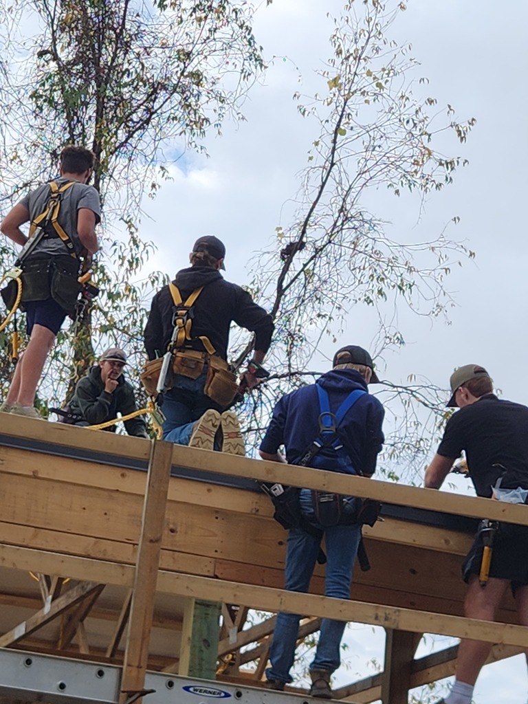A group of young men wearing hard hats work on and around the frame of a large barn.