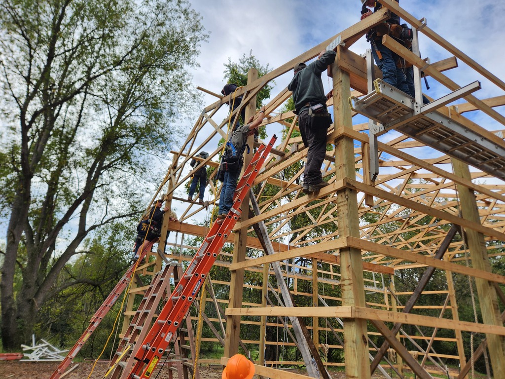 A group of young men wearing hard hats work on and around the frame of a large barn.