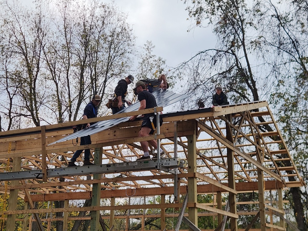 A group of young men wearing hard hats work on and around the frame of a large barn.