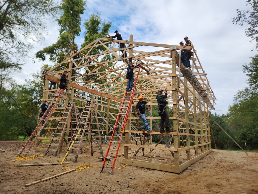 A group of young men wearing hard hats work on and around the frame of a large barn.