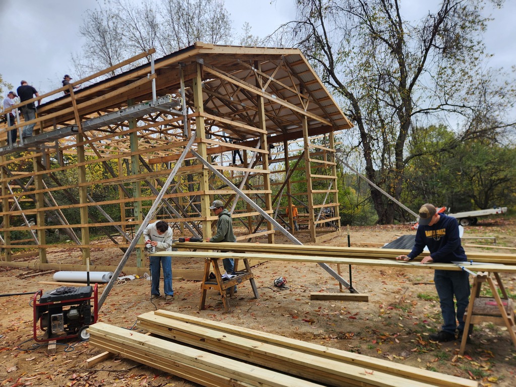 A group of young men wearing hard hats work on and around the frame of a large barn.