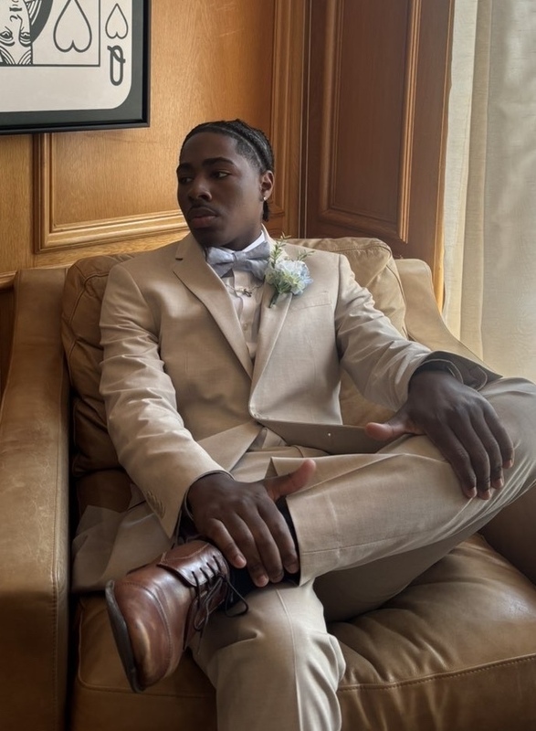 High school male student seated in a chair with a tan suit on and his legs folded as he looks off camera