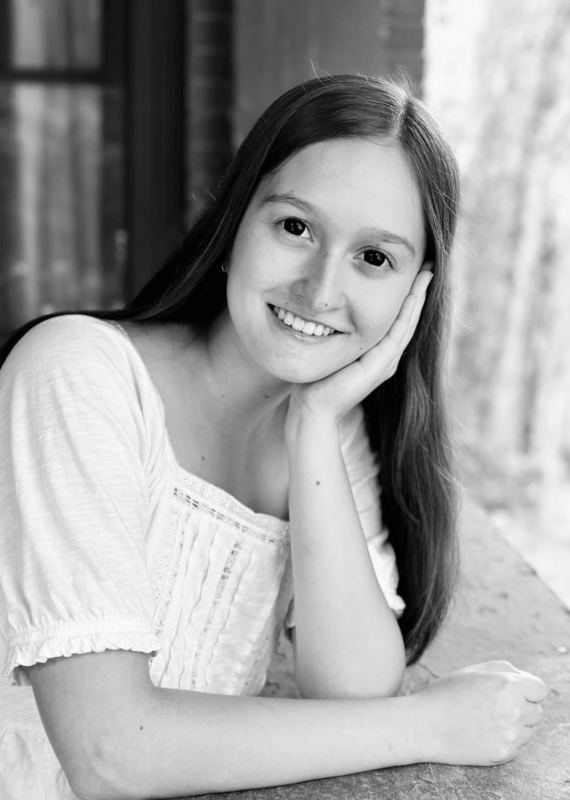 Black and white photo of a female high school student who is resting her cheek on her hand and smiling at the camera.