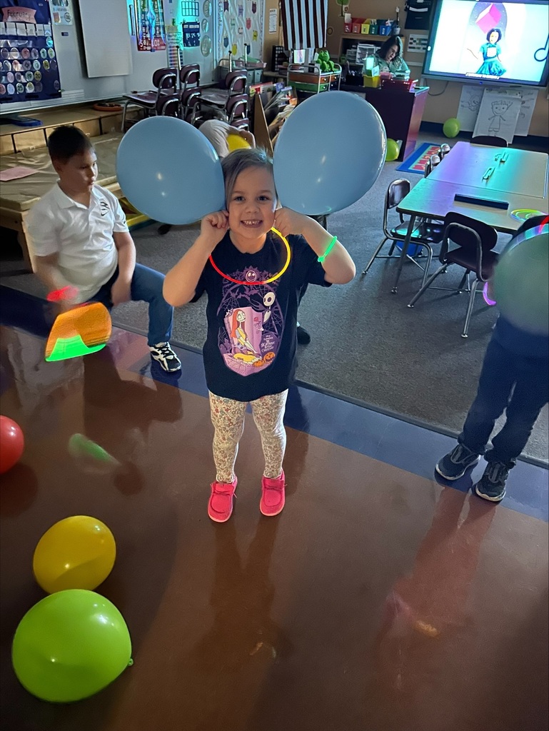 A child stands in a room with balloons, wearing blue balloons on her ears. Two people sit nearby, and tables with chairs are in the background.