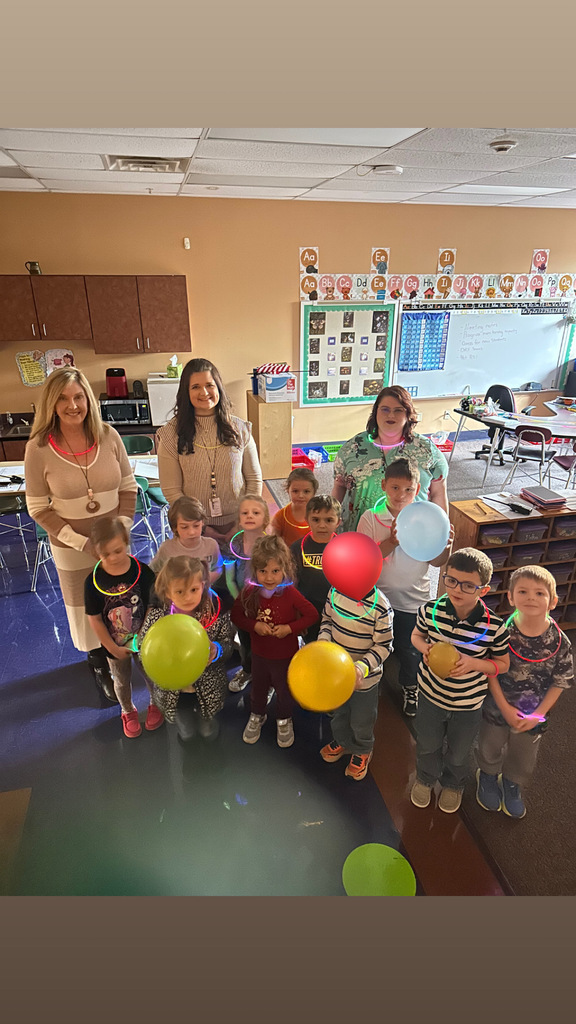 Group of children and adults standing in a classroom, smiling. Several children hold balloons. Behind them, a bulletin board, white board, and tables.