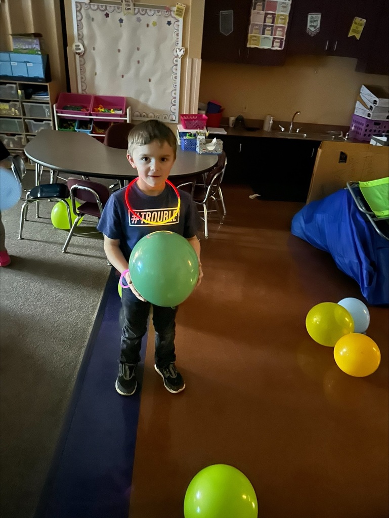 A boy holds a green balloon in a room with a table, chairs, balloons, and a blue rug.