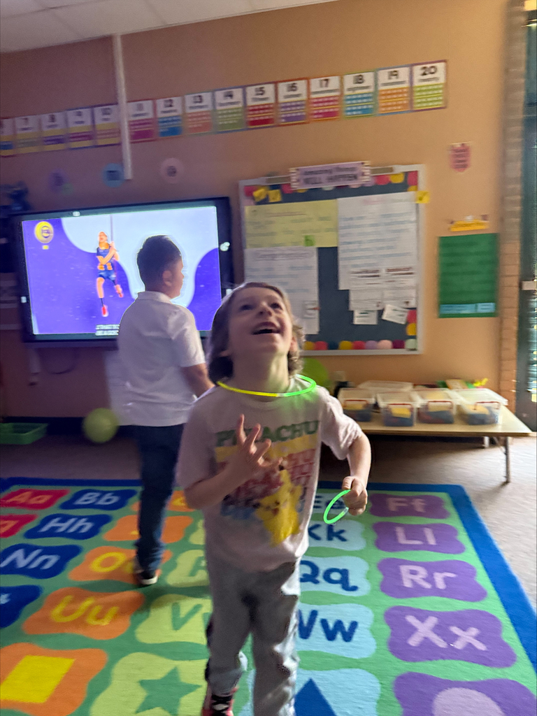 Two children in a classroom. One with glowing headband and holding glowsticks laughs. A large screen and alphabet rug.