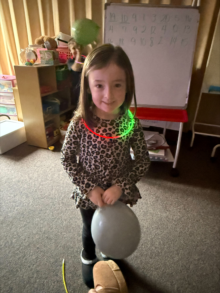 A girl with a lit-up necklace stands in a room, holding a balloon. Behind her, a whiteboard displays letters and numbers. A wooden shelf with toys and books is to the left.