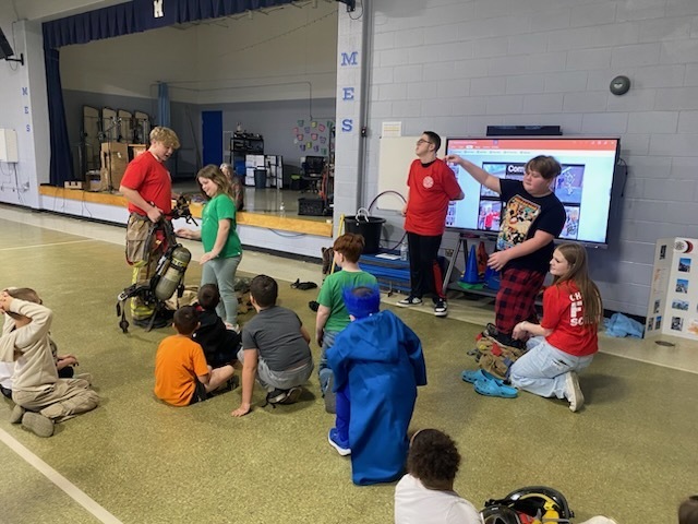 A classroom with children seated on the floor and standing. A man in a red shirt gestures toward a large screen.