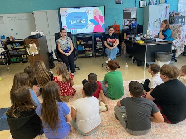 Children sit in a circle around two women in a classroom, with a large screen and shelves in the background.