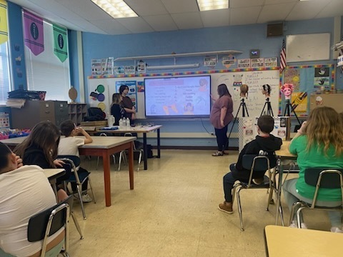 A classroom with students seated in chairs. Two individuals stand in front of a projector screen.