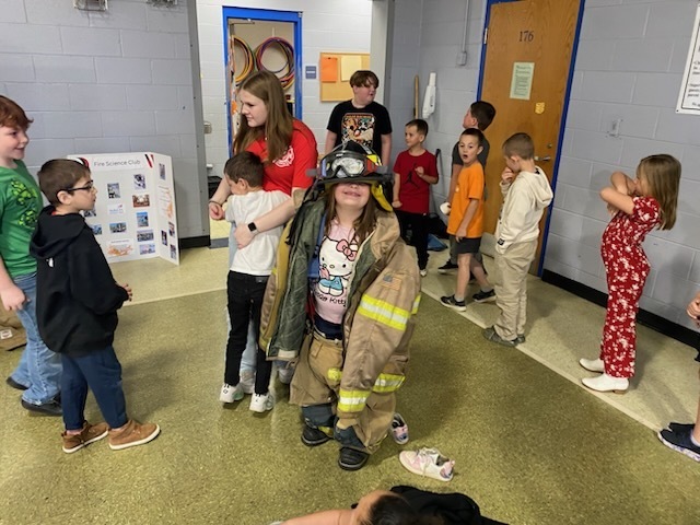 Kids in a room, one dressed as a firefighter, with a woman and others watching.