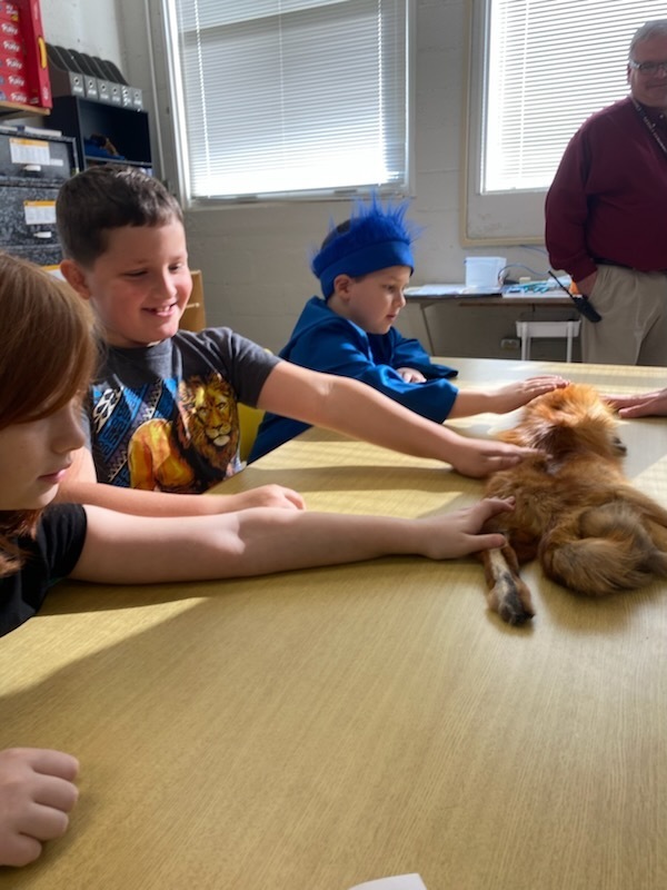 Children seated at a table with a small dog. One child pets the dog. A man stands nearby.