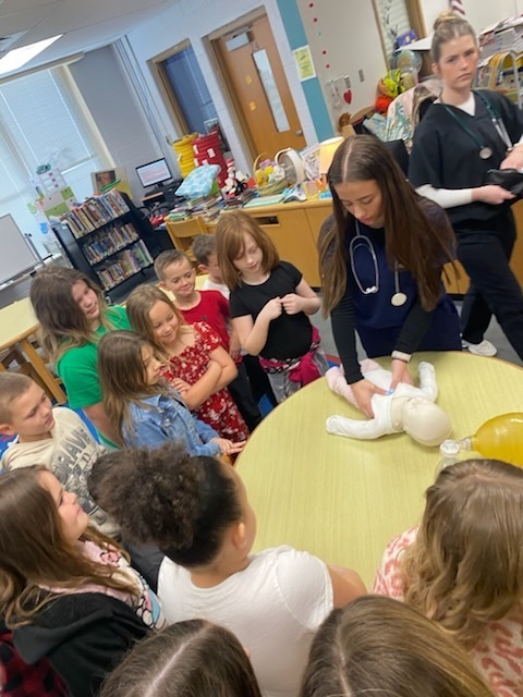 Several children gather around a table, watching a woman in blue scrubs demonstrate CPR on a dummy.