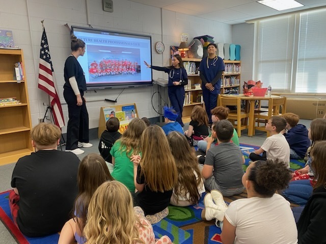 Several children sit in a circle on a rug, listening to two adults standing in front of a screen.