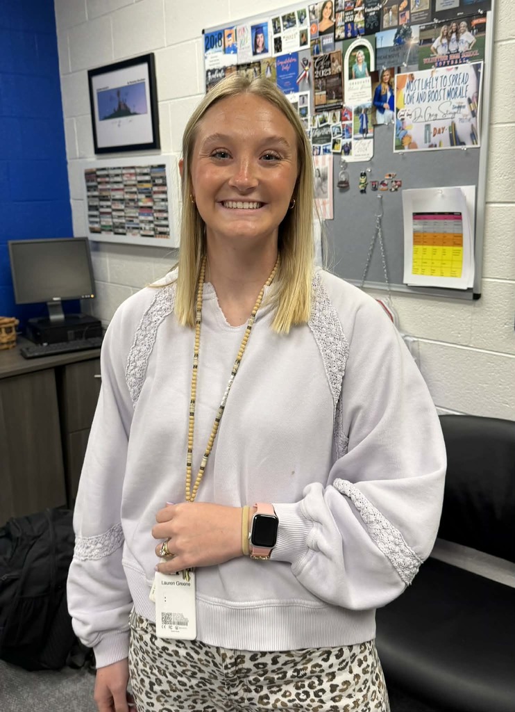 A woman with blonde hair in a classroom with a blue wall, wearing a white sweater, leopard print pants, and a necklace.