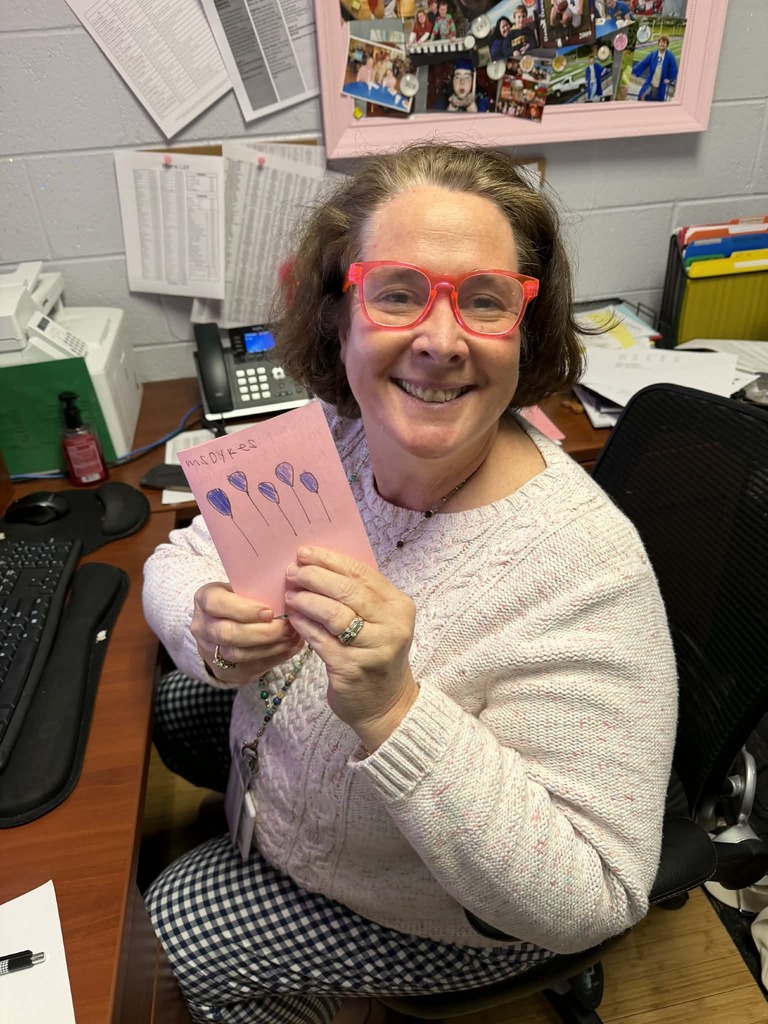 Woman in glasses and a white sweater holds a pink card, smiling, seated at a desk.