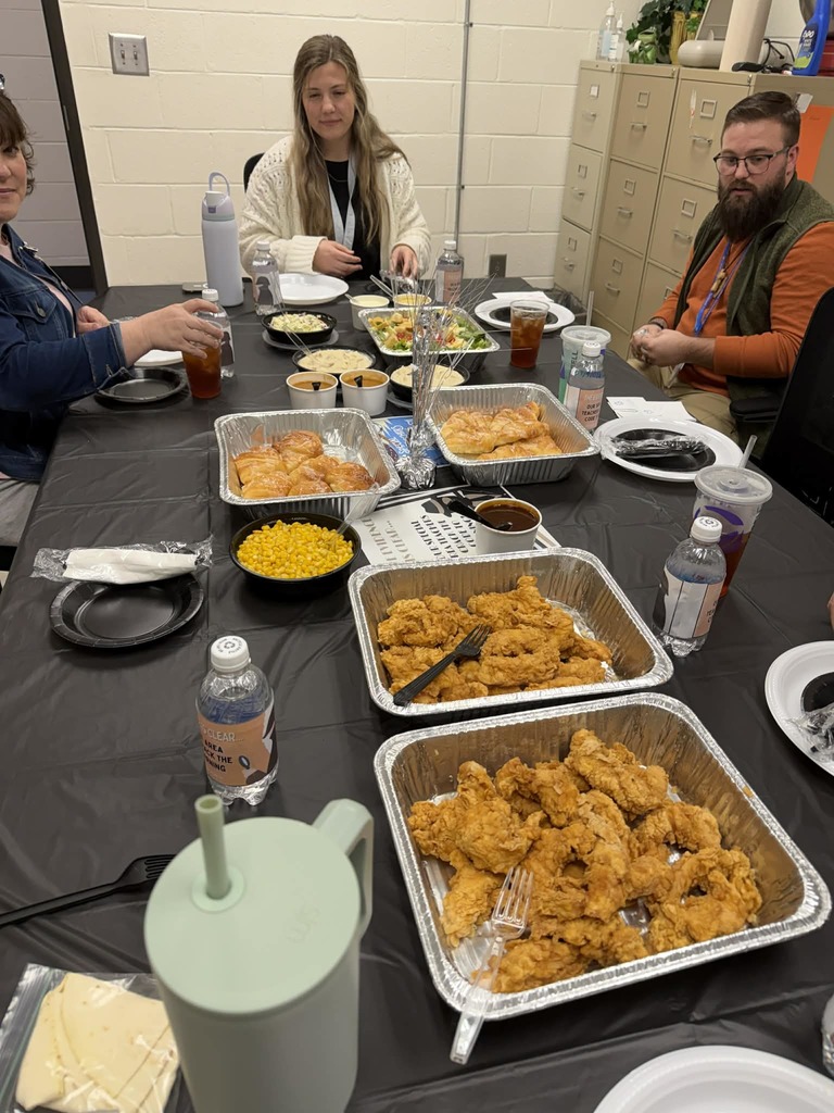 People seated at a long table with various foods in trays and bowls, including fried chicken, corn, and a salad.