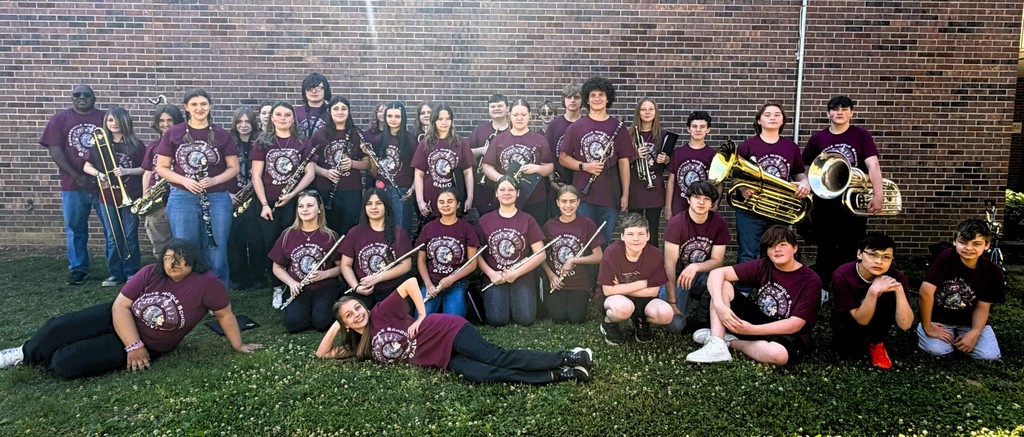 A group of young people with band instruments in matching maroon shirts pose on a grassy area in front of a brick building.