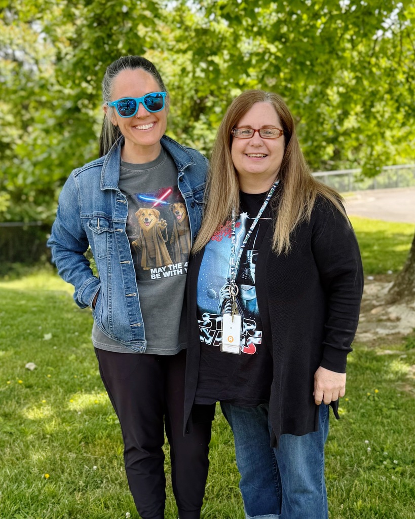 Two women stand in a grassy area with trees in the background, smiling at the camera.
