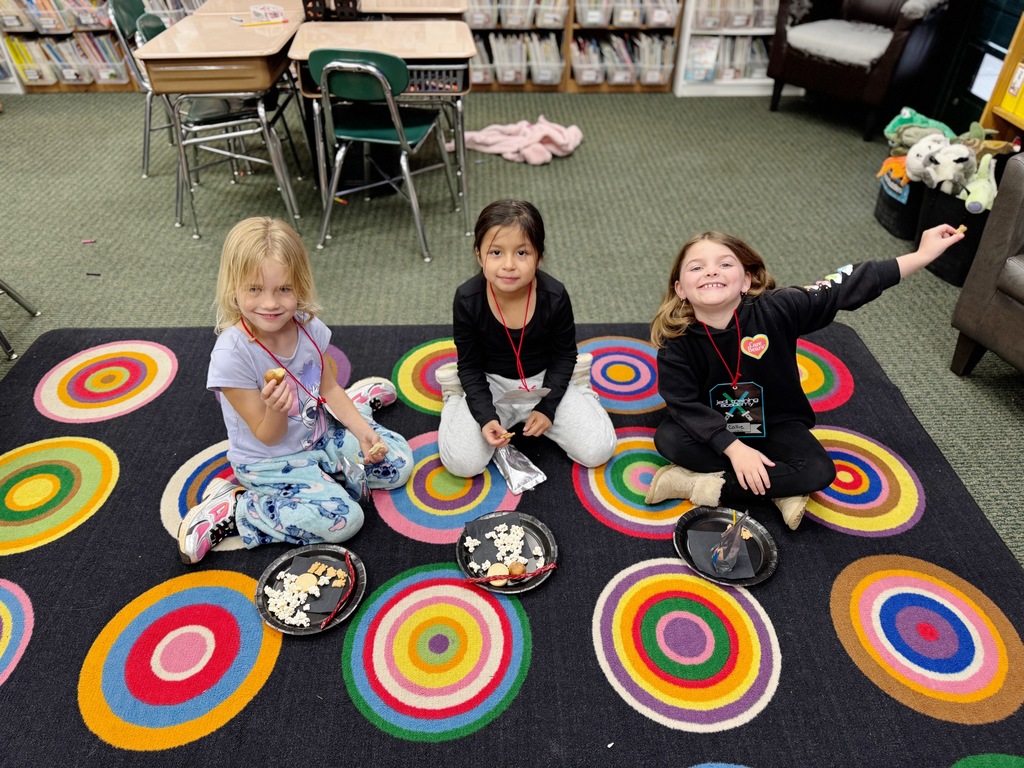 Four young girls sit on a colorful floor mat in a classroom. Each holds a plate of food.