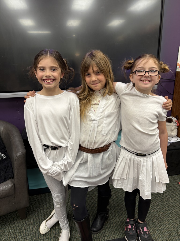 Three girls in white outfits with belts and boots, smiling, stand in a room with a large screen.