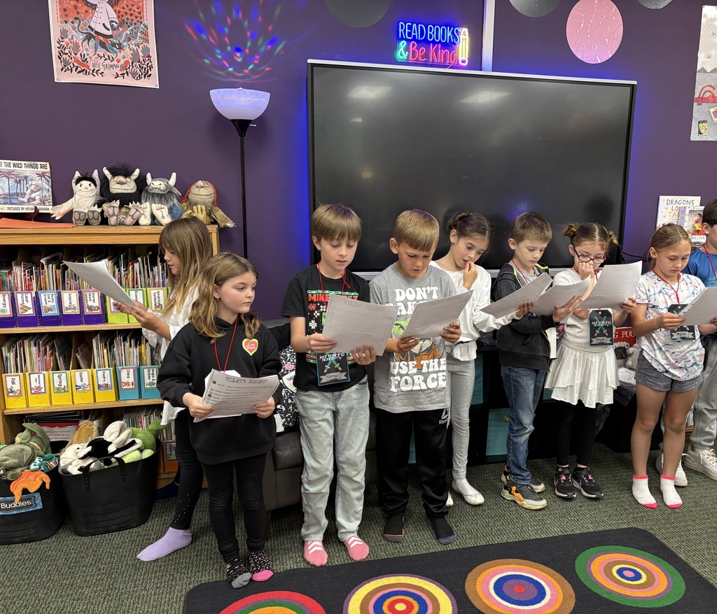 Nine children stand in a classroom, holding papers and reading. A large monitor is behind them, and shelves with books are to the left.