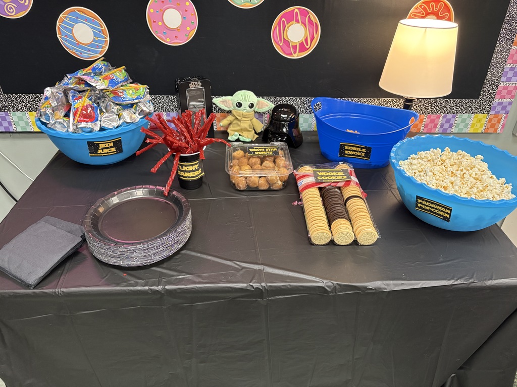 A table covered with a black tablecloth has snacks, bowls, and a lamp. A blackboard with colorful circles is in the background.