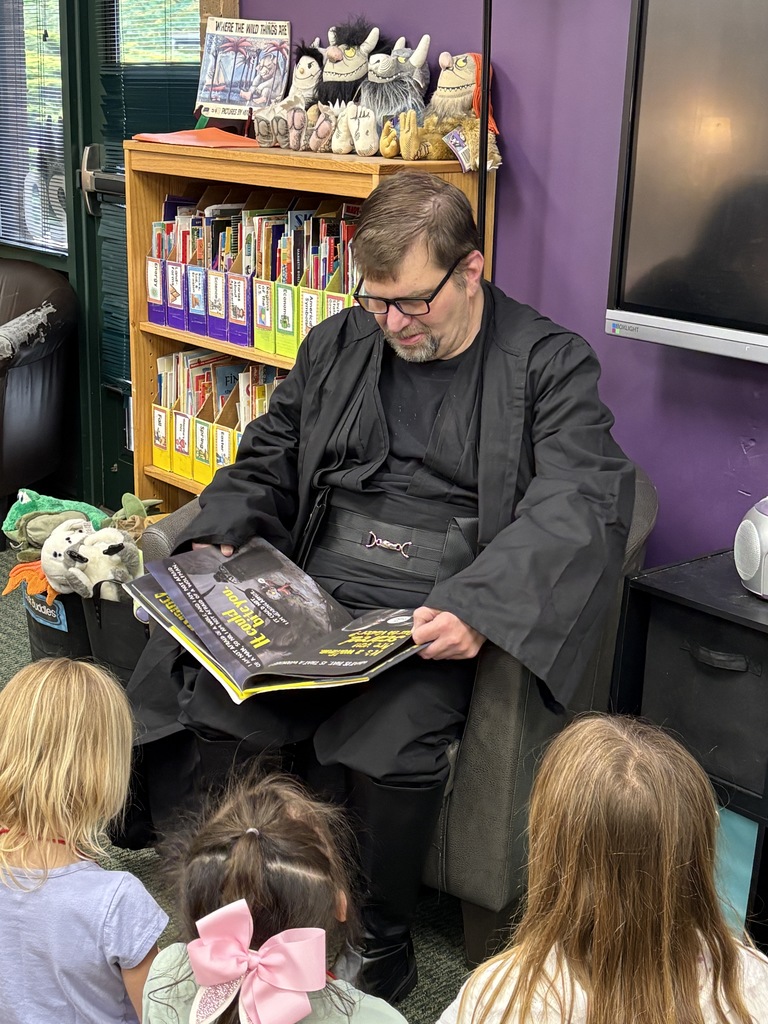 Man in a black robe reads a comic book to children sitting on the floor in a library.