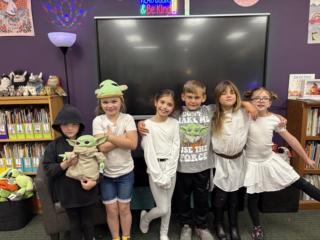 A group of children stand together in a room with shelves of books. One holds a plush toy.