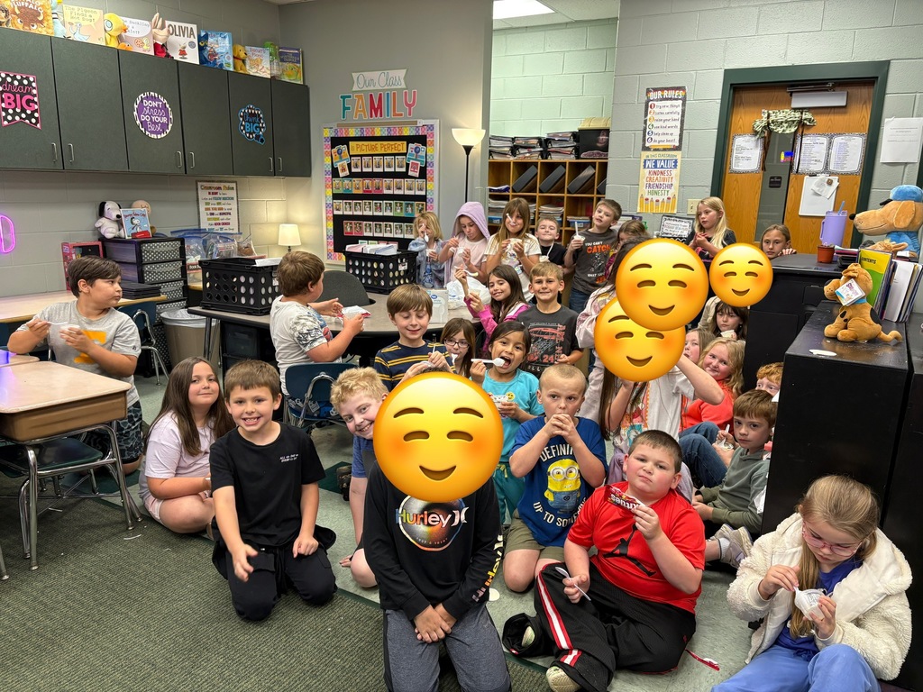 Children sit on the floor holding emoji balloons. Some wear casual clothes. Behind, tables, chairs, shelves, and a lamp.