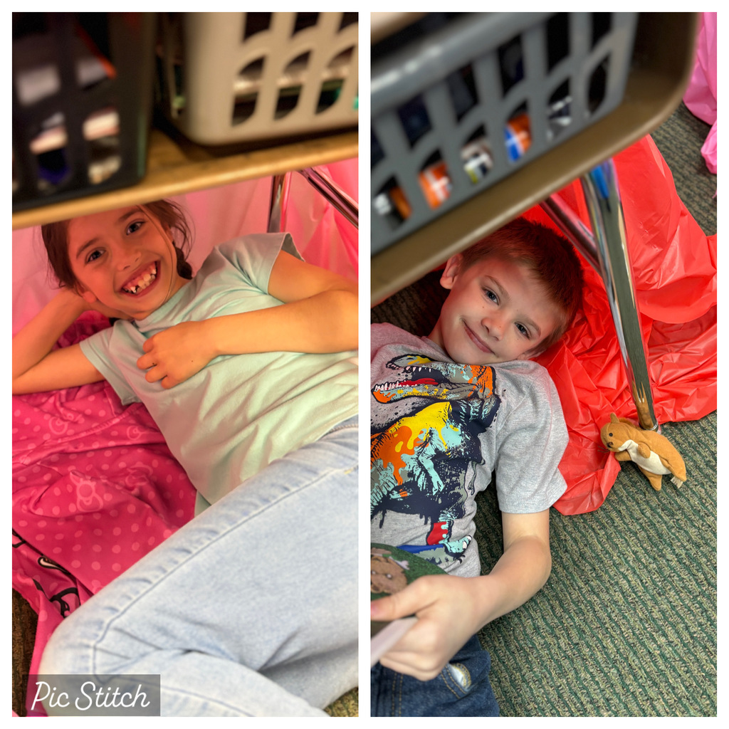 Two children in a classroom, one lying on a bed, the other under a desk, both smiling.