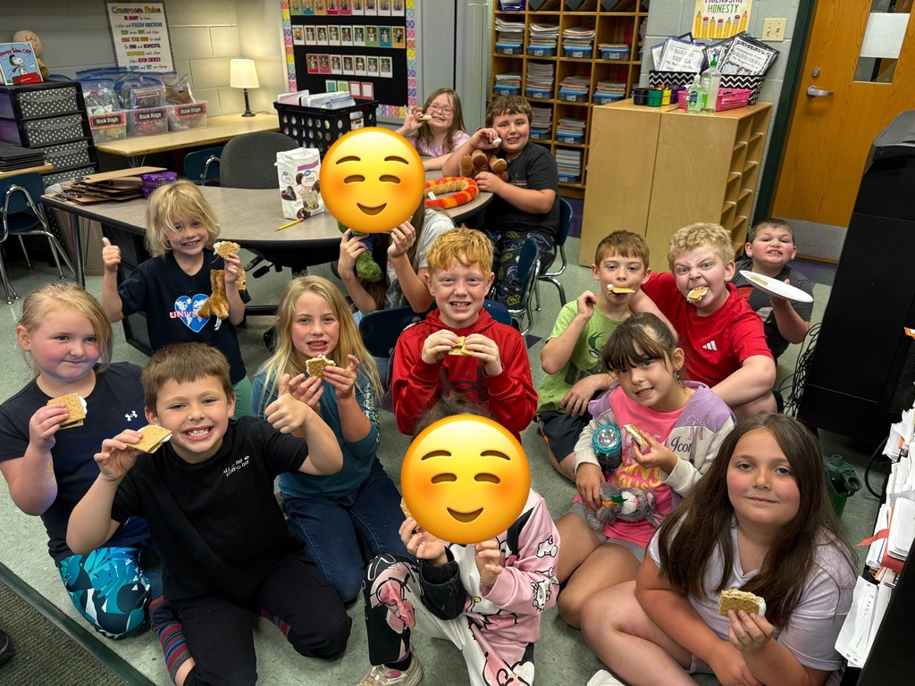 A group of children sits on the floor in a classroom, holding plates of food and smiling. Some have emojis on their plates.