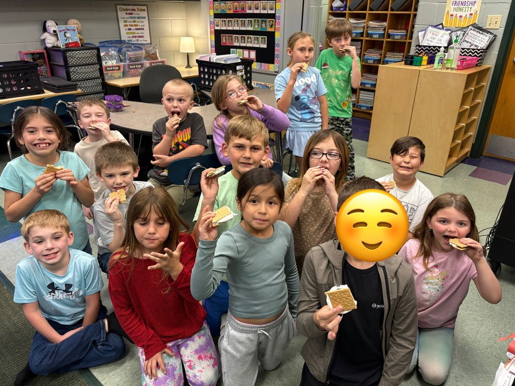 Several children in a classroom, some holding food, posing for a group photo. They sit on the floor.
