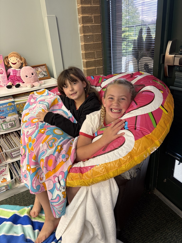 Two children sit in a bean bag chair holding a pink floater. Behind them is a shelf with books and toys.