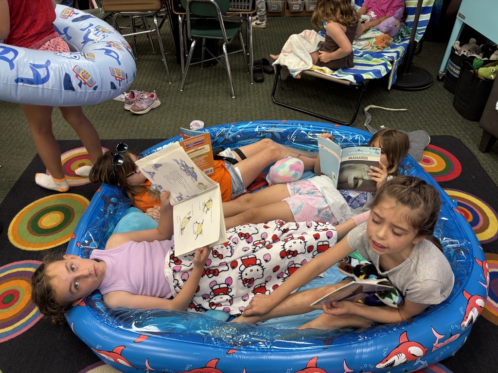 Four children sit in an inflatable pool, reading books. Two girls lie down while the other two sit upright.
